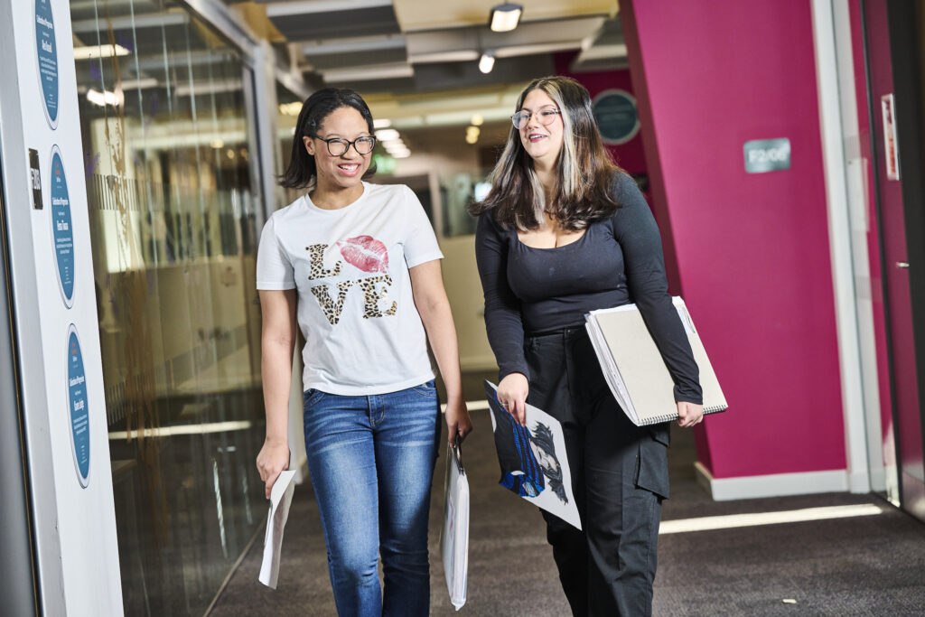 Two students walking down a corridor with their work in their hands